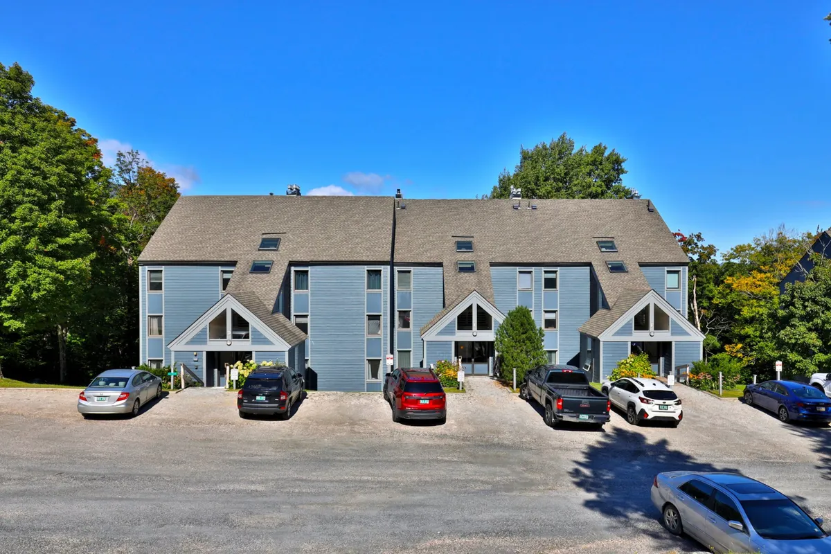 A view of the multi-unit building enveloped by trees, with a parking area out front and a sense of community. The scene suggests easy arrival and access to both the property and natural surroundings, hinting at the shuttle service and group experiences.