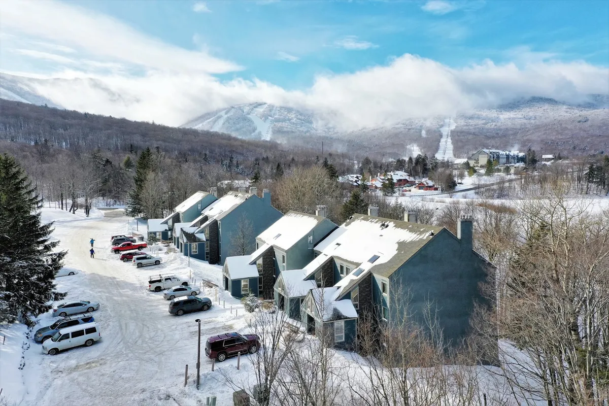 A snowy exterior view of the buildings set against a mountain backdrop, with people skiing nearby and parked cars, capturing the winter atmosphere and the excitement of ski-home access.