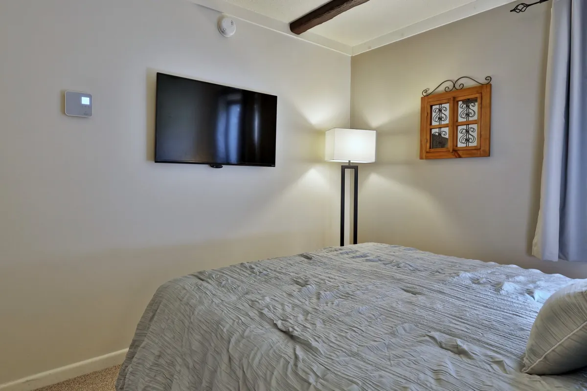A cozy bedroom with a textured bedspread, a flat-screen TV, a standing lamp, and stylish wall art. The ambiance is calming, designed for unwinding with entertainment or a good book before bed.