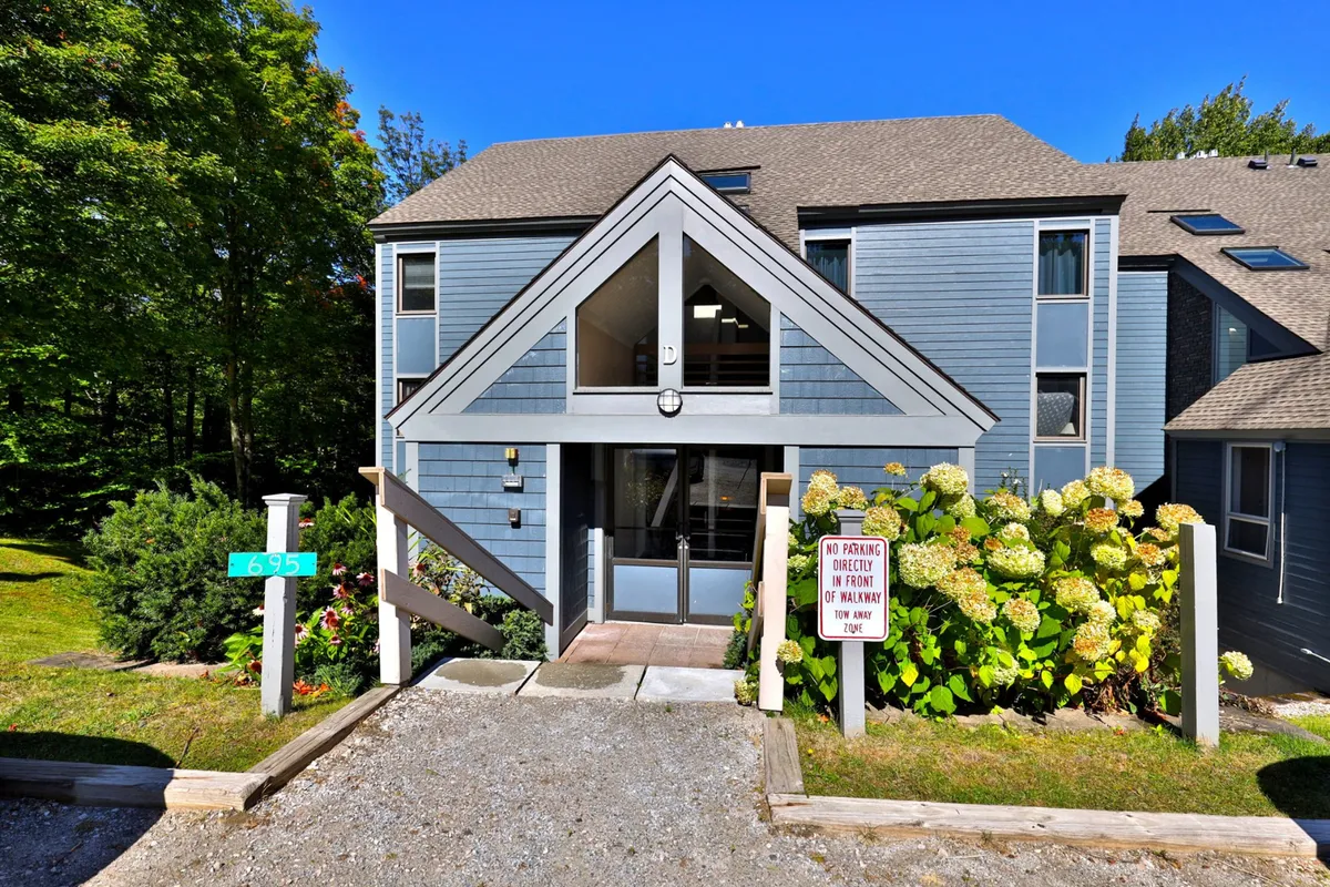The image features the entrance structure of the condo complex, surrounded by a landscaped garden, a house number sign, and a visible parking sign. The space looks welcoming and is well-maintained, evoking a sense of arrival and community.