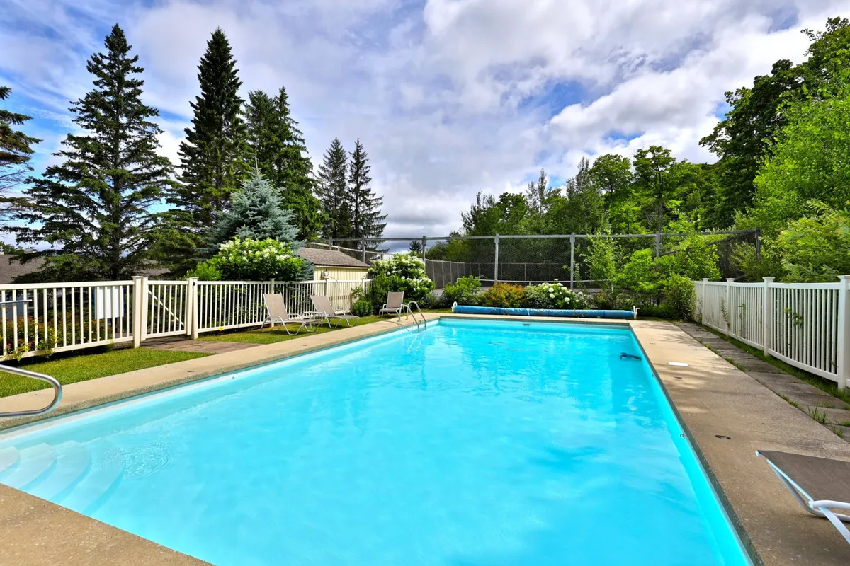 This image features a sparkling outdoor swimming pool surrounded by lush greenery and enclosed by a fence. The sun casts a warm glow on the water, and there is ample space for lounging and socializing around the pool.