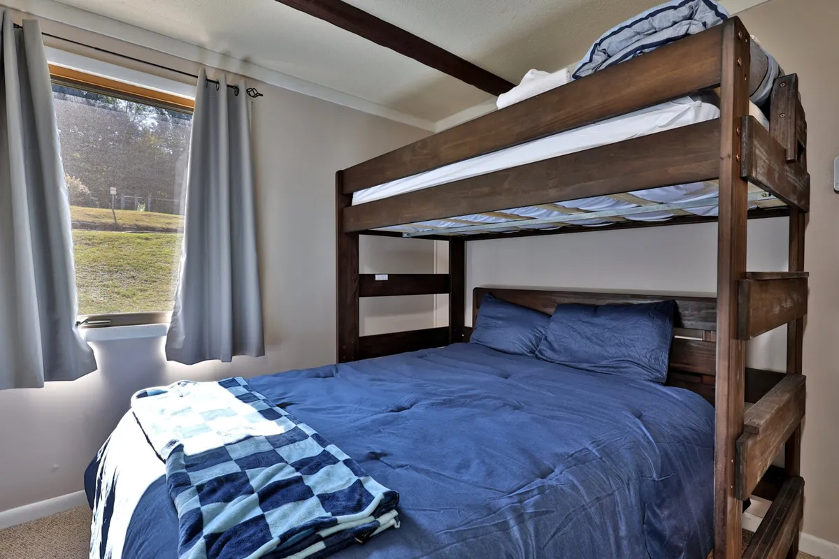 A cheerful bunk room with a twin-over-queen bunk bed dressed in blue bedding. The space is filled with natural light from a nearby window, making it both lively and functional for families or groups. A sturdy ladder provides safe access to the upper bunk.