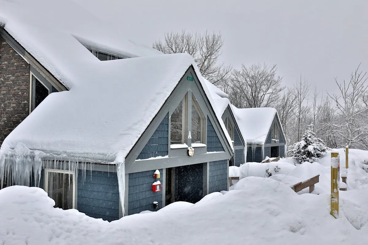 A tranquil exterior shot of snow-covered rooftops, icicles hanging from wooden railings, and snow-dusted trees surrounding the property. The scene evokes a cozy winter retreat ambiance, suggestive of returning home to warmth after a day outdoors.